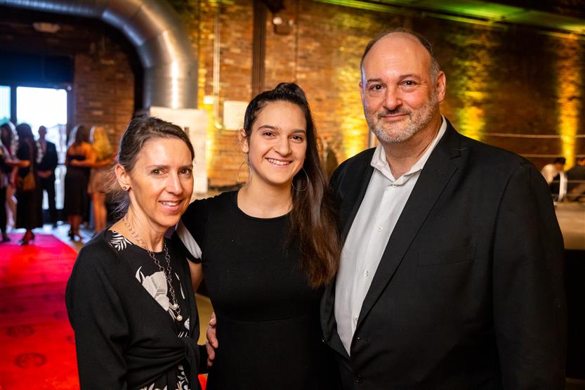 Two women, both wearing black dresses, smile with a man wearing a black blazer